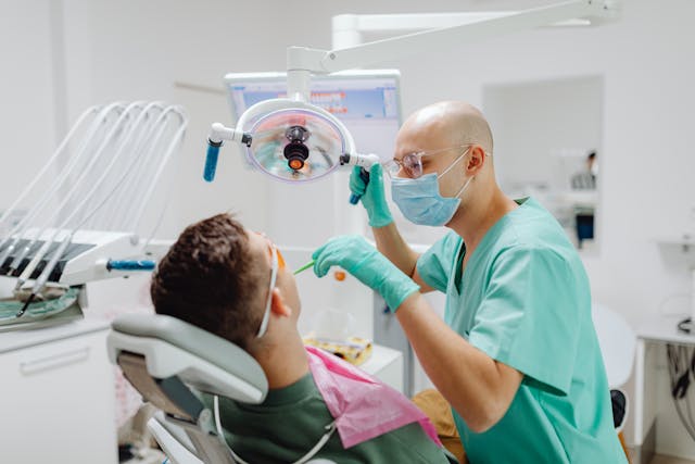 A dentist wearing a mask and teal scrubs adjusts a surgical light while examining a patient’s teeth in a modern dental clinic.